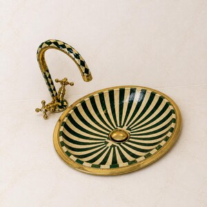 May include: An oval bathroom sink featuring a green and cream striped pattern with a gold-toned rim and drain. A matching curved faucet with a green and cream diamond pattern and gold-toned cross handles is positioned next to the sink basin.
