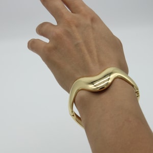 Può includere: Un bracciale rigido incernierato in metallo color oro lucido con un design liscio e ondulato. La superficie metallica presenta una finitura altamente riflettente che si curva attorno al polso, fissata con un discreto meccanismo a cerniera.
