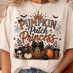 May include: A cream-colored t-shirt featuring a graphic print with the text PUMPKIN PATCH PRINCESS. The design includes a crown, bats, spiderwebs, a skull, various pumpkins with bows, and a coffee cup, all in orange, black, and leopard print.