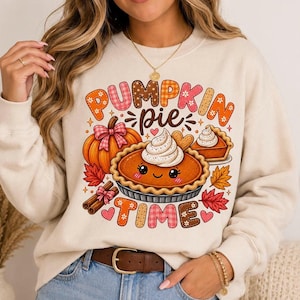 Puede incluir: Una sudadera de cuello redondo color crema con un estampado gráfico que incluye el texto PUMPKIN PIE TIME. El diseño muestra un pastel de calabaza sonriente con nata montada, una calabaza con un lazo rosa, ramas de canela y hojas de otoño.