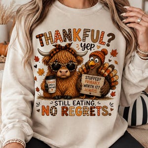 Puede incluir: Una sudadera beige de cuello redondo con un gráfico de una vaca Highland con gafas de sol y un pavo con gorro de punto. El texto dice THANKFUL? YEP. STILL EATING. NO REGRETS. El pavo sostiene un cartel que dice STUFFED? PROBABLY. WORTH IT? ALWAYS.