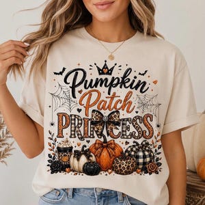 May include: A cream-colored t-shirt featuring a graphic print with the text Pumpkin Patch Princess. The design includes illustrations of pumpkins, a leopard print bow, spiderwebs, bats, and a coffee cup, all in orange, black, and white.