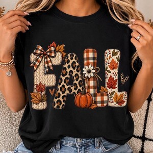May include: A black t-shirt featuring the word FALL in large letters. The F has a plaid bow and knit texture, the A has a leopard print, the L has a plaid pattern with a daisy and pumpkin, and the second L features autumn leaves and berries.