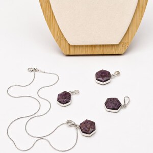 May include: A jewelry set consisting of a silver chain necklace and three matching pendants. Each pendant features a faceted, hexagonal, deep purple stone set in a polished silver frame. The necklace includes a delicate silver chain with a clasp.