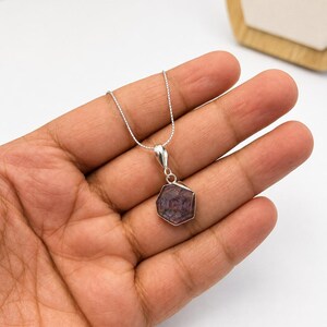 May include: A silver necklace featuring a hexagonal pendant with a dark purple and reddish-brown gemstone. The stone displays a natural star-like pattern within its faceted surface, held by a simple silver bail on a delicate silver box chain.