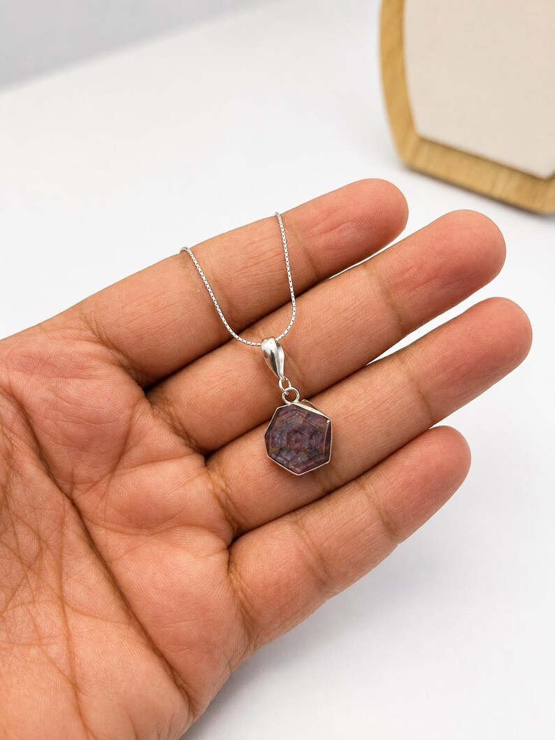 May include: A silver necklace featuring a hexagonal pendant with a dark purple and reddish-brown gemstone. The stone displays a natural star-like pattern within its faceted surface, held by a simple silver bail on a delicate silver box chain.