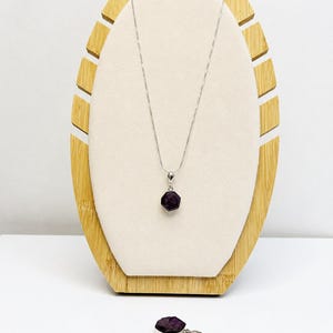 May include: A silver necklace featuring a hexagonal dark purple gemstone pendant, accompanied by three matching hexagonal dark purple gemstone charms with silver loops, displayed on a light-colored oval stand with a bamboo-style frame.