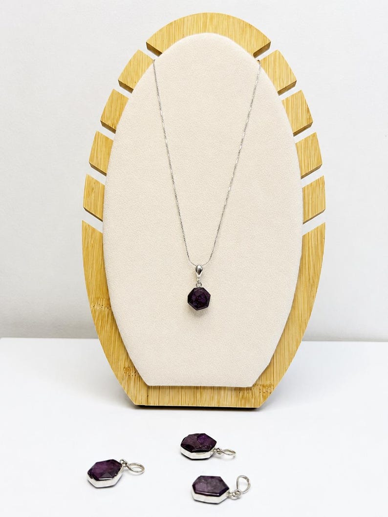 May include: A silver necklace featuring a hexagonal dark purple gemstone pendant, accompanied by three matching hexagonal dark purple gemstone charms with silver loops, displayed on a light-colored oval stand with a bamboo-style frame.