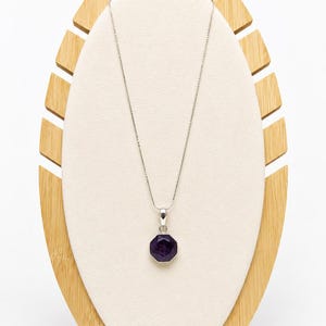 May include: A silver necklace featuring a thin box chain and a faceted, octagonal deep purple gemstone pendant. The pendant is held by a simple silver bail, and the entire piece is displayed on a light-colored, oval-shaped jewelry stand.