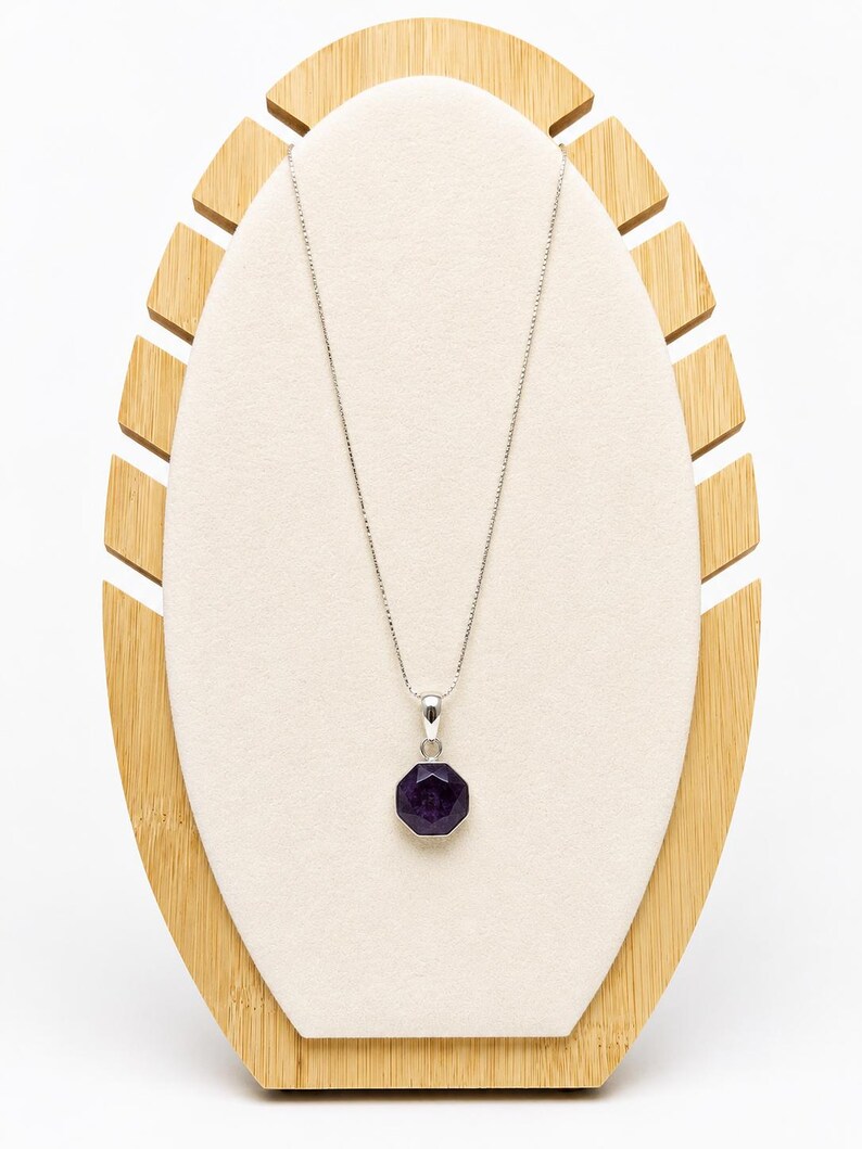 May include: A silver necklace featuring a thin box chain and a faceted, octagonal deep purple gemstone pendant. The pendant is held by a simple silver bail, and the entire piece is displayed on a light-colored, oval-shaped jewelry stand.