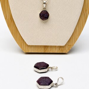 May include: A silver necklace featuring a hexagonal dark purple gemstone pendant, accompanied by two matching hexagonal dark purple gemstone pendants with silver bails, each displaying a faceted surface and a polished metallic setting.