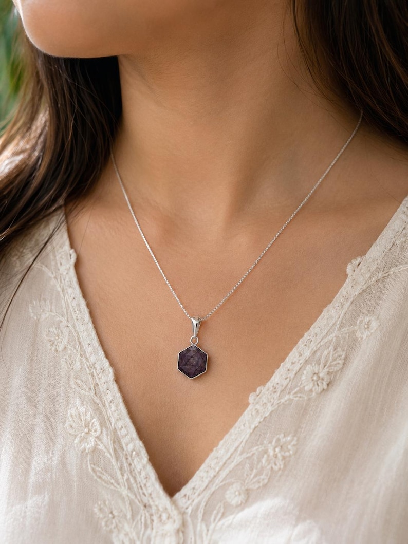 May include: A silver necklace featuring a hexagonal pendant with a dark purple, textured stone inlay. The pendant is suspended from a delicate silver chain, showcasing a clean, geometric design that rests against the collarbone area.