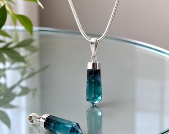 May include: A silver-toned snake chain necklace featuring a faceted, hexagonal, deep blue and teal crystal pendant. A second, identical crystal pendant lies horizontally on the surface, showcasing the same polished, pointed gemstone design.
