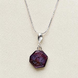 May include: A silver box chain necklace featuring a hexagonal, deep reddish-purple gemstone pendant. The stone displays natural internal banding and a polished surface, suspended from a simple silver bail attached to the delicate metal chain.