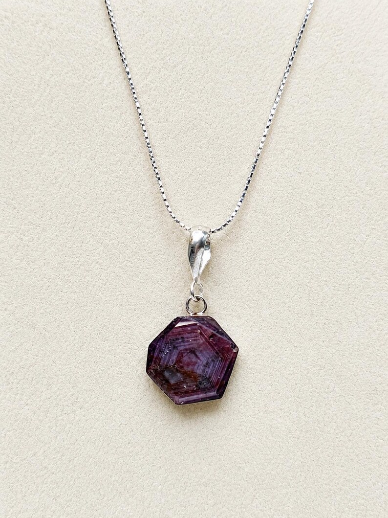May include: A silver box chain necklace featuring a hexagonal, deep reddish-purple gemstone pendant. The stone displays natural internal banding and a polished surface, suspended from a simple silver bail attached to the delicate metal chain.