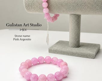 May include: Two bracelets made of smooth, round, light pink beads labeled as Pink Argonite. One bracelet is displayed on a gray T-bar stand, and the other rests on a flat surface. Text on the image reads Gulistan Art Studio and Stone name Pink Argonite.
