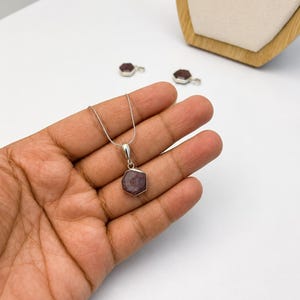 May include: A silver necklace featuring a hexagonal pendant with a dark reddish-purple gemstone. The pendant is suspended from a thin silver chain. Two additional matching hexagonal gemstone pendants are visible in the background.