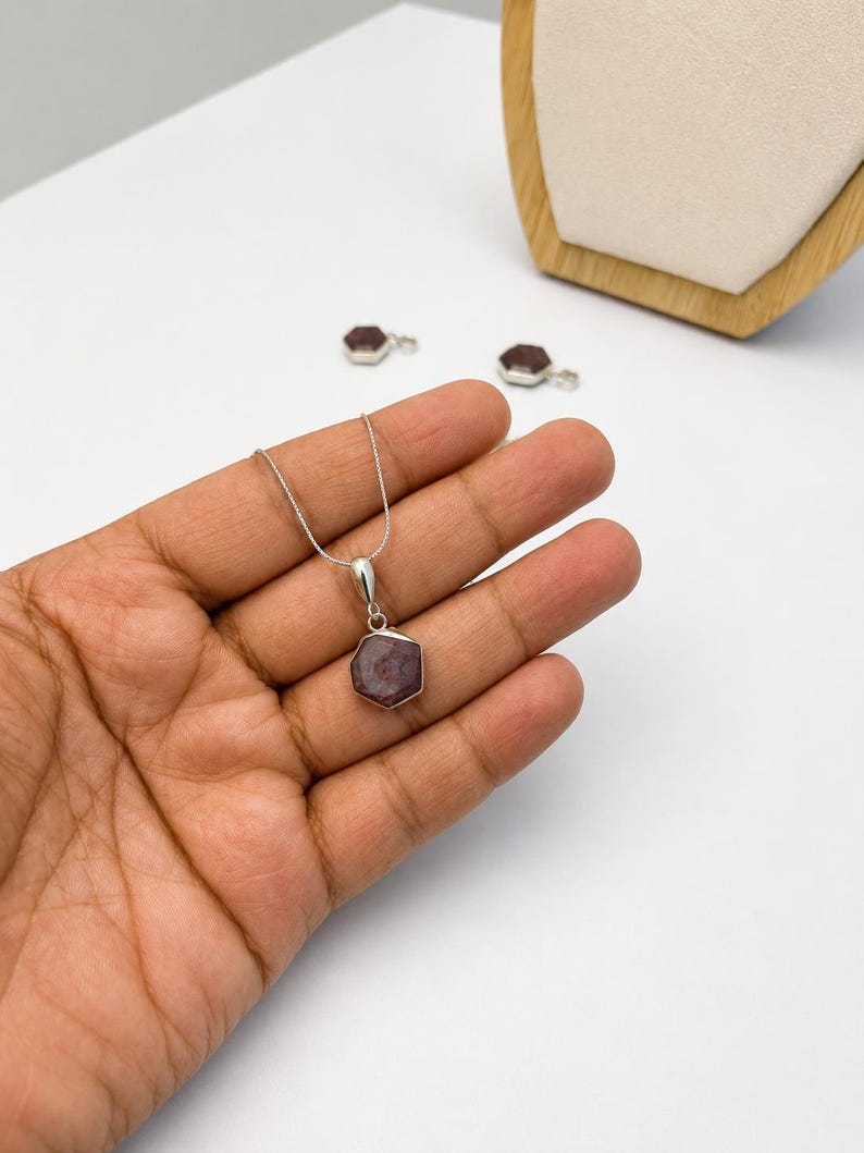 May include: A silver necklace featuring a hexagonal pendant with a dark reddish-purple gemstone. The pendant is suspended from a thin silver chain. Two additional matching hexagonal gemstone pendants are visible in the background.