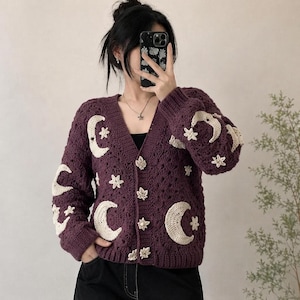 May include: A deep purple crochet cardigan featuring cream-colored crescent moon and star appliques. The garment has a V-neckline, long sleeves, and a front button closure with four distinct leaf-shaped buttons made of a light-colored material.