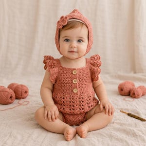 May include: A dusty rose crochet romper featuring ruffled sleeves, a square neckline, and three wooden buttons down the front. The outfit includes a matching pointed bonnet adorned with a crochet flower detail on the side, crafted from yarn.