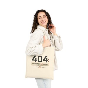 May include: A cream-colored canvas tote bag featuring the black text 404: MOTIVATION NOT FOUND printed on the front. Below the text is a small graphic of a warning triangle symbol with two lightning bolts on either side of the icon.