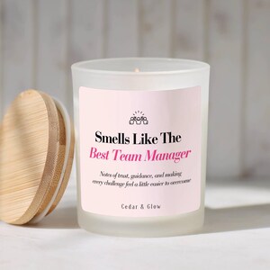 May include: A frosted glass candle jar with a light pink label featuring the text Smells Like The Best Team Manager, Notes of trust, guidance, and making every challenge feel a little easier to overcome, and Cedar & Glow, accompanied by a round bamboo lid.