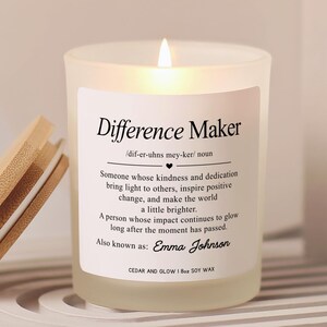 May include: A frosted glass candle in a jar with a lit wick and a bamboo lid. The white label features the text Difference Maker, a dictionary-style definition, and the name Emma Johnson, with the scent listed as Cedar and Glow 8 oz soy wax.