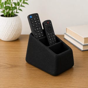 May include: A black, textured remote control holder with two rectangular compartments. Two black remote controls with various buttons and icons are placed inside the slots. The holder has a slanted front profile and a solid, matte finish.