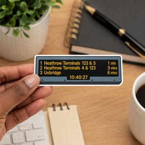 Op de afbeelding: Een rechthoekige sticker met een digitaal vertrekbord in de stijl van de Londense metro met gele tekst. Het toont: 1 Heathrow Terminals 123 & 5 1 min, 2 Heathrow Terminals 4 & 123 3 mins, 3 Uxbridge 6 mins, en de tijd 10:40:27.