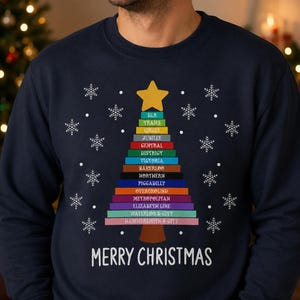 May include: A navy blue crewneck sweatshirt featuring a graphic of a Christmas tree constructed from horizontal colored bars representing London Underground lines, topped with a yellow star, white snowflakes, and the text MERRY CHRISTMAS.