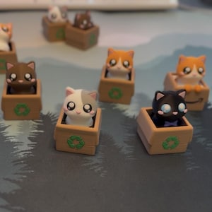 May include: A collection of small, stylized cat figurines, each sitting inside a miniature cardboard box. The cats feature various colors including orange, black, white, and brown. Some boxes display a green recycling symbol or a black smile logo.