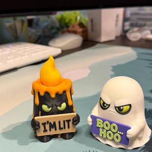 May include: Two small figurines: a black candle with an orange flame holding a tan sign that reads I'M LIT, and a white ghost with green eyes holding a purple sign that reads BOO... HOO. Both figures have angry expressions and are made of plastic.