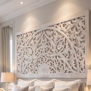 May include: A large, rectangular, off-white wooden headboard featuring intricate, symmetrical, hand-carved floral and scrollwork patterns. The headboard is mounted on the wall above a bed dressed in white linens and a light beige throw blanket.