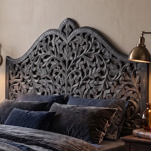 May include: A dark charcoal grey wooden headboard featuring an intricate, hand-carved floral and leaf pattern with a curved, arched top. The headboard is positioned behind a bed dressed in dark blue and grey velvet bedding and pillows.