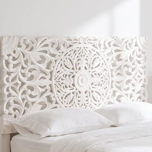 Può includere: Una testiera in legno bianco caratterizzata da un intricato motivo floreale e circolare a mandala intagliato a mano. La testiera è posizionata dietro una struttura letto con biancheria bianca, inclusi due cuscini e un piumino, per un look luminoso.