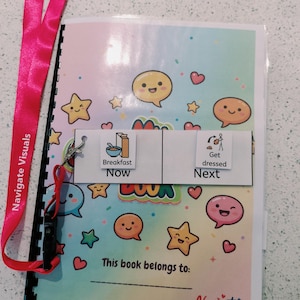 May include: A spiral-bound visual schedule book with a pastel gradient cover featuring cartoon stars and speech bubbles. It includes a bright pink lanyard labelled "Navigate Visuals" and two removable cards showing "Breakfast Now" and "Get dressed Next".