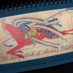 May include: A spiral-bound notebook featuring a colorful illustration of a Native American warrior on horseback. The artwork, in shades of red, blue, and yellow, is set against a grid-lined background. The text "The Ledgerbook of Thomas Blue-Eagle" is written in cursive.