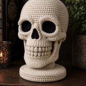 Puede incluir: Una escultura de cráneo tejida a ganchillo con hilo color hueso, que presenta cuencas oculares negras profundas y una cavidad nasal negra. El cráneo muestra una mandíbula detallada con dientes individuales y descansa sobre una base circular.