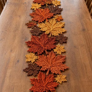May include: A table runner composed of numerous crocheted maple leaves in various sizes and shades of autumn colours, including deep brown, burnt orange, golden yellow, and rust red, arranged in a long, dense, and overlapping linear pattern.