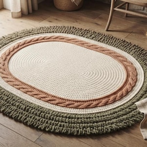 May include: An oval-shaped area rug featuring a cream-colored center, a textured terracotta-colored braided border, and a wide outer edge made of thick, dark olive green yarn loops. The rug has a layered, multi-textured design with distinct sections.
