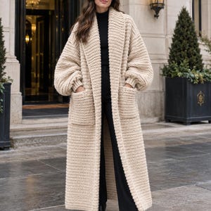 May include: A long, beige, open-front knit cardigan featuring a thick ribbed texture, wide lapels, dropped shoulders, full-length sleeves with ribbed cuffs, and two large patch pockets on the front, worn over a black turtleneck and black trousers.