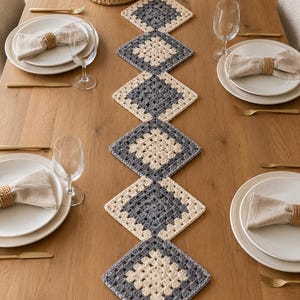Op de afbeelding: Een gehaakte tafelloper bestaande uit zes verbonden, ruitvormige granny squares in afwisselend crème en donkergrijs garen. De loper ligt in het midden van een houten eettafel die is gedekt met witte borden en servetten.
