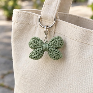 May include: A sage green crocheted butterfly charm attached to a silver-toned metal lobster clasp and ring. The butterfly features a textured, knotted yarn construction with a central wrapped detail, hanging from a plain off-white canvas tote bag.