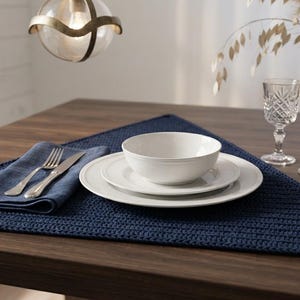 May include: A dark blue triangular crocheted placemat featuring a textured stitch pattern. The placemat is set with a white ceramic dinner plate, a smaller salad plate, a white bowl, a folded dark blue cloth napkin, a silver fork, and a knife.