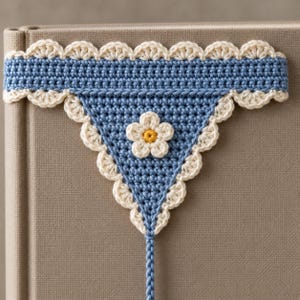 May include: A triangular crochet bookmark featuring a blue base with a cream-coloured scalloped border. A small, five-petalled cream flower with a yellow centre is attached to the centre of the triangle, and a thin blue braided cord extends downwards.
