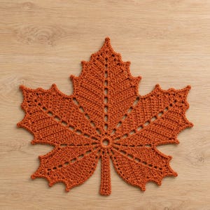 May include: A burnt orange crochet doily shaped like a maple leaf. The textile features a central circular opening with radiating openwork patterns and intricate stitching that defines the pointed lobes and serrated edges of the leaf design.