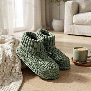 May include: A pair of sage green ankle-high slippers crafted from thick, chunky knit yarn. The slippers feature a prominent ribbed cuff at the top and a textured, woven construction throughout the body, providing a soft and substantial appearance.