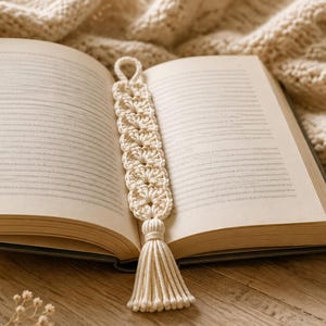 Peut inclure: Un marque-page en crochet blanc cassé présentant un motif floral répétitif, une petite boucle sur le dessus et un long pompon épais à la base. Le marque-page est posé verticalement au centre d'un livre ouvert aux pages couleur crème.