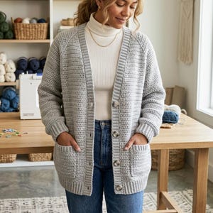 May include: A light gray, long-sleeved, crocheted cardigan featuring a ribbed collar, cuffs, and hem. The garment has a button-down front with four visible buttons and two large patch pockets on the front. It is worn over a white turtleneck.