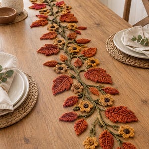 Puede incluir: Un camino de mesa de ganchillo que presenta una enredadera central verde con hojas texturizadas de color naranja, rojo y marrón, junto con pequeñas flores amarillas de centro oscuro. El camino está dispuesto en un patrón sinuoso sobre la mesa.